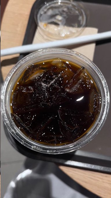 A close-up overhead shot shows a clear plastic cup filled with dark iced coffee and ice cubes, sitting on a tray next to another cup, a white straw, and chopsticks.