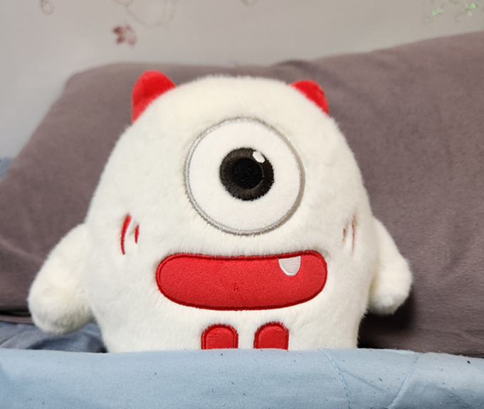 A fluffy white plush toy with one large eye, red horns, a red mouth with a single fang, and small red feet, resting on a blue cushion against a grey pillow.
