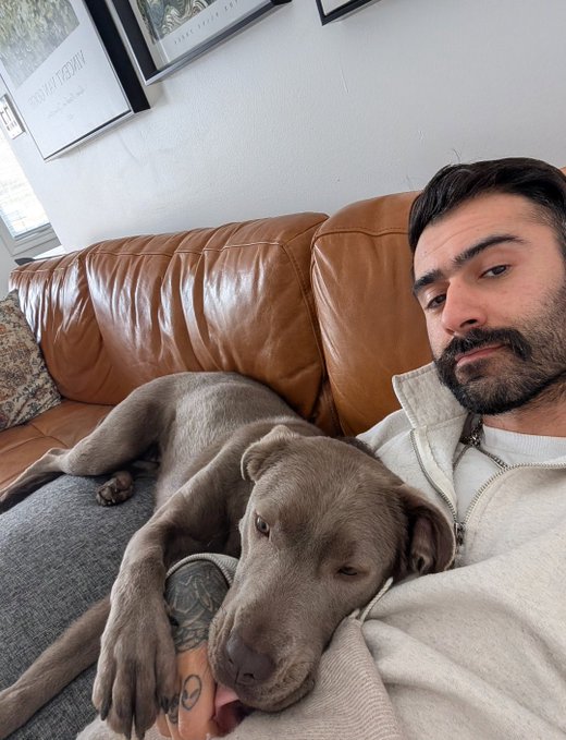 The image shows a man sitting on a couch with his dog resting its head on him. The man has tattoos on his arm, and there are pictures hanging on the wall behind them. The photo captures a moment of relaxation between the man and his dog in their living room.