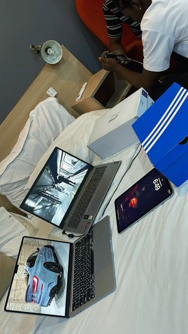 Two open laptops and a smartphone are displayed on a white bed, alongside an Adidas shoebox and a white drawstring bag, while a person sits in the background using their phone.