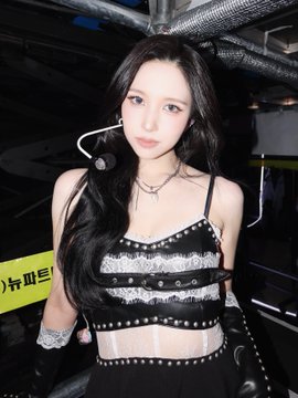 A young woman with long dark hair poses backstage in a black studded crop top and arm warmers, with Korean text visible on yellow tape behind her, while the caption "MINA OH MY GOD" suggests the post is celebrating her appearance.