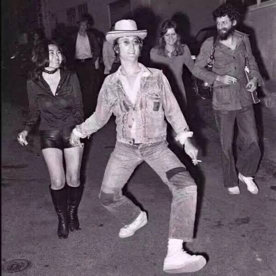 The image features a group of people, including two women and one man, all dressed up for an event or gathering. The woman on the left side of the photo appears to be dancing with her friends while holding hands, wearing black boots that match the overall style of the scene. On the right side of the photo, there is another person who seems to be smoking a cigarette. The group's attire and activities suggest they are attending an event or social gathering where everyone is dressed up for a special occasion.