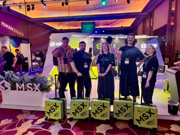 A group of five people stand on a stage at an MSX event, posing behind branded green MSX boxes, with a 'TOKEN 2049' banner visible in the background.