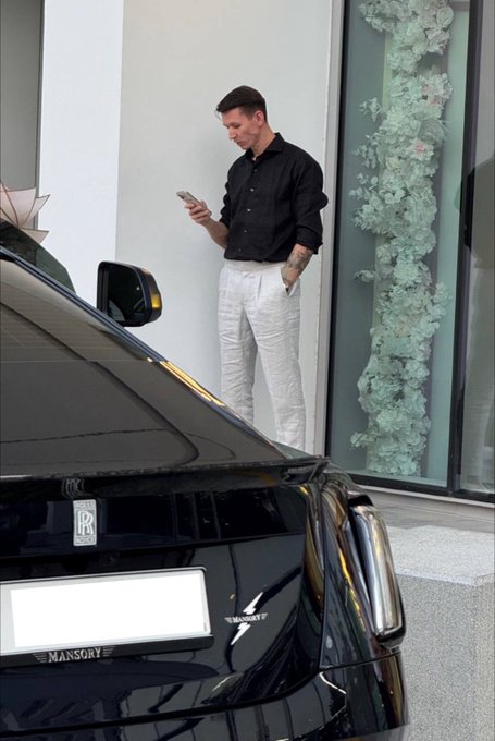 A well-dressed man in a black shirt and white pants checks his phone while standing next to a black Rolls-Royce Mansory. The luxury scene contrasts with the post's skeptical question about NFTs making a "legendary comeback," highlighting the disconnect between traditional symbols of wealth and the speculative crypto market.
