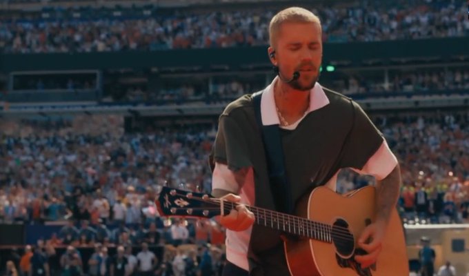 A man with a buzzcut plays an acoustic guitar in a packed stadium while wearing a headset microphone. The image complements the post's tribute by depicting a large-scale, emotionally charged performance befitting the "GOAT" acclaim.