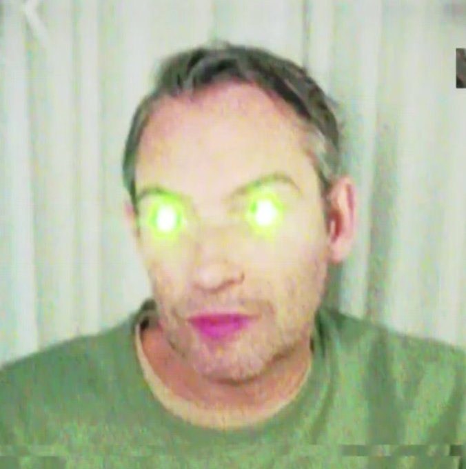 A blurry webcam photo of a person with digitally altered glowing green eyes and purple lips, giving an unsettling appearance.