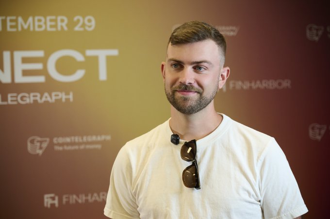 A bearded man wearing a white t-shirt with sunglasses hanging from the collar poses in front of a step-and-repeat backdrop featuring "CONNECT," "COINTELEGRAPH," and "FINHARBOR" branding, corresponding to the event where GenSyn's Ben Fielding discussed how AI will expand trading beyond assets to include predictions and beliefs.