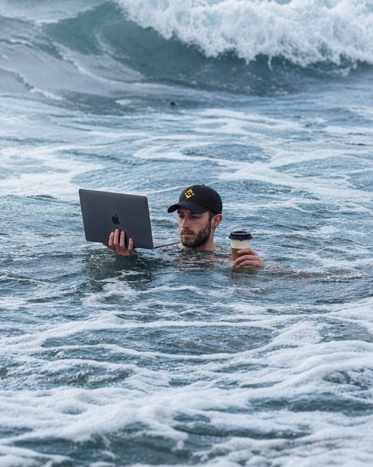 A man submerged in ocean waves works on an Apple laptop while holding a takeaway coffee cup, embodying the post's message "you can leave the desk, not the market" by humorously showing dedication to staying connected with the financial markets even from the surf, with his black cap displaying "CA" lettering visible amid the churning whitewater.