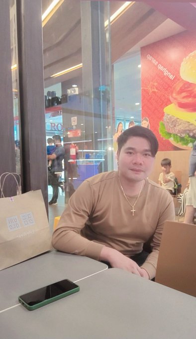 A man wearing a tan long-sleeve shirt and cross necklace sitting at a fast-food restaurant table next to a Uniqlo shopping bag.