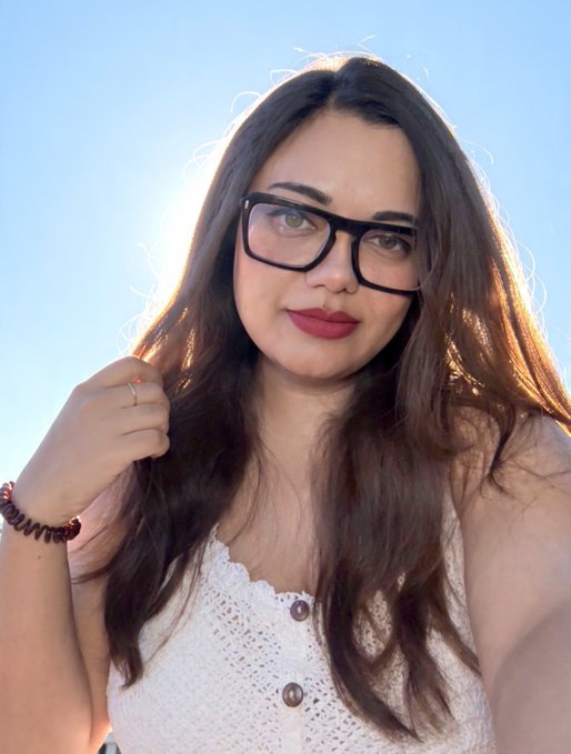 A woman with long dark hair and glasses wears a white top while posing outdoors against a bright blue sky, wearing a beaded bracelet and rings.