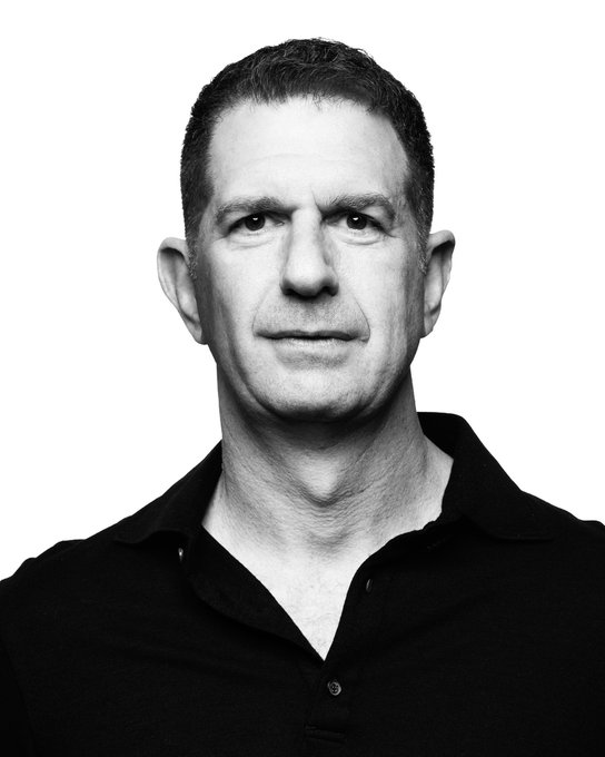 A black and white portrait photograph of a middle-aged man with short hair wearing a black polo shirt against a plain background.