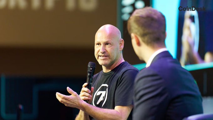 The image shows two men sitting next to each other at a table with microphones, one of whom appears to be speaking into a microphone while the other listens attentively. The man speaking has a bald head and is wearing a black t-shirt that says "ETH" on it. He seems to be discussing the potential of Ethereum as a fully ZK-proof protocol for atomic execution and unified liquidity across different layers, without the need for bridges or middlemen.