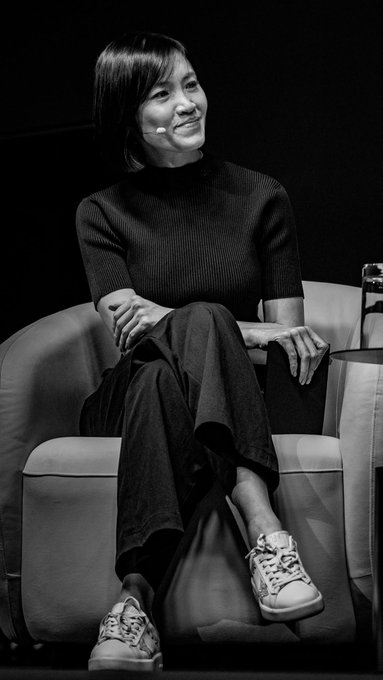 A woman with a short bob haircut sits on a stage in a black outfit and patterned sneakers, speaking at what appears to be a panel discussion. The image complements the post about VIP traders discussing AI, resilient execution, and crypto market dynamics, suggesting she is one of the featured speakers (likely @thatismyquant, @azkeepsimple, or @vctbtc) sharing insights with the audience.