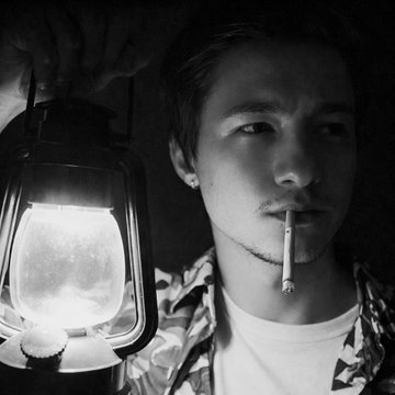 A black and white photo of a young man smoking a cigarette, holding up an illuminated lantern in a dark setting.