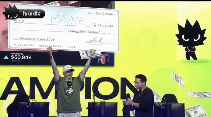 A stage event showing a giant check made out to 'MAYNE' for $25,000.00 from 'hudi' dated Oct 7, 2026, with a man holding up the check while another holds a microphone.