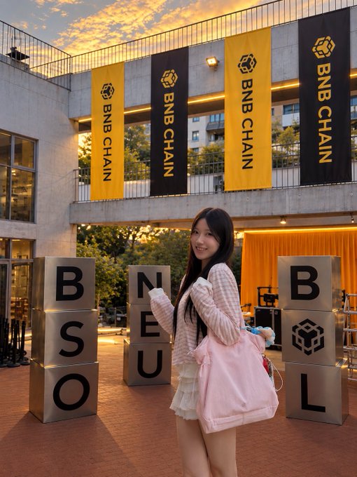 A photograph of a smiling young woman with long dark hair in a pink cardigan and skirt, posing at a BNB Chain/BNB Seoul event with branded yellow banners and cube displays in the background.