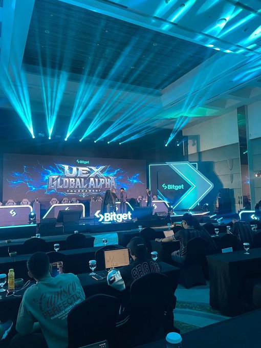 A Bitget "UEX GLOBAL ALPHA TOURNAMENT" event with blue stage lighting and laser effects, featuring a large screen and arrow-shaped displays, with attendees seated at tables watching the presentation.
