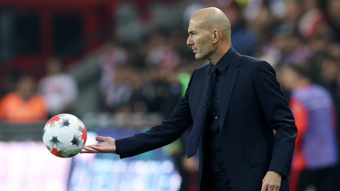 A bald man in a dark suit and tie on a soccer field, holding a white and red soccer ball in his hand.