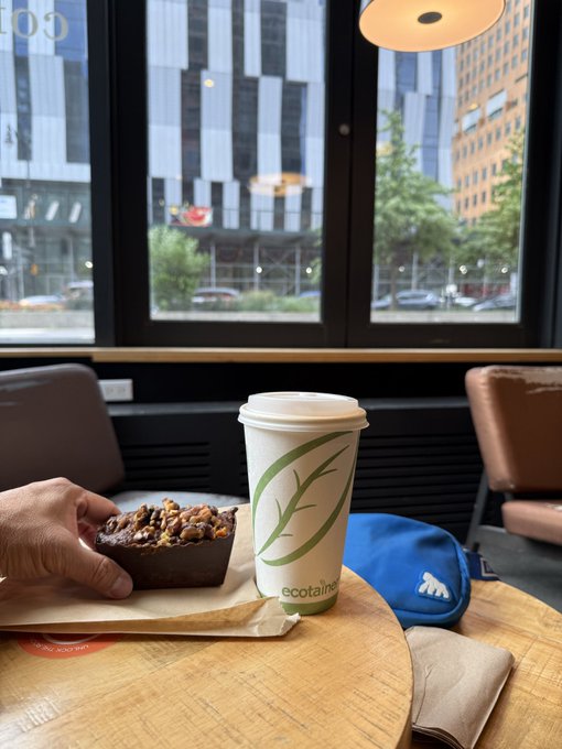 A person's hand reaches for a chocolate nut pastry next to an 'ecotainer' branded coffee cup on a round wooden table inside a cafe with a city view through the window.