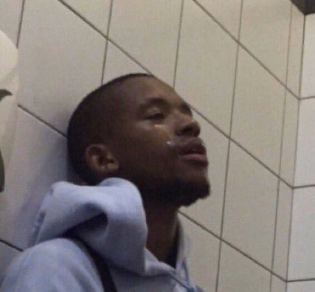 A man in a grey hoodie leaning against a white tiled wall with his eyes closed.