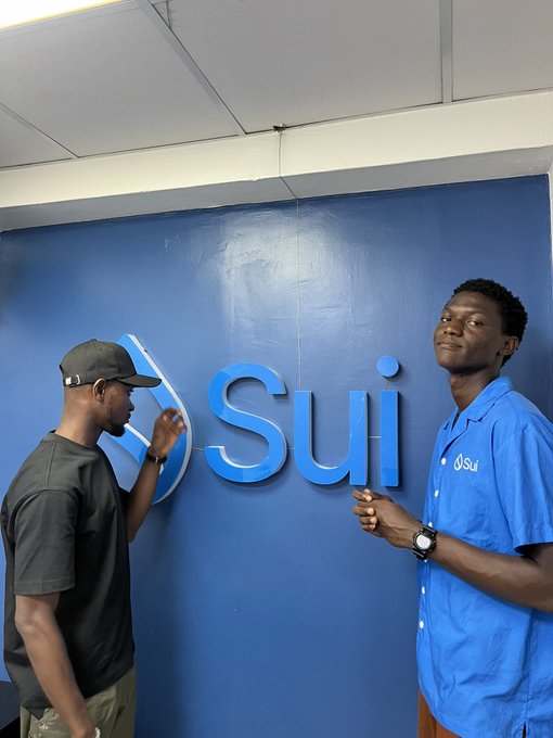 The image shows two men standing next to each other against a blue wall with the word "Sui" written on it. One of them is holding a pen while the other one has his hand up, possibly pointing at something or making a gesture. The background appears to be a room where they are located.