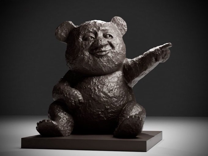 A bronze sculpture of a smiling teddy bear sitting on a base with its arm outstretched, displayed in a gallery with dramatic lighting.
