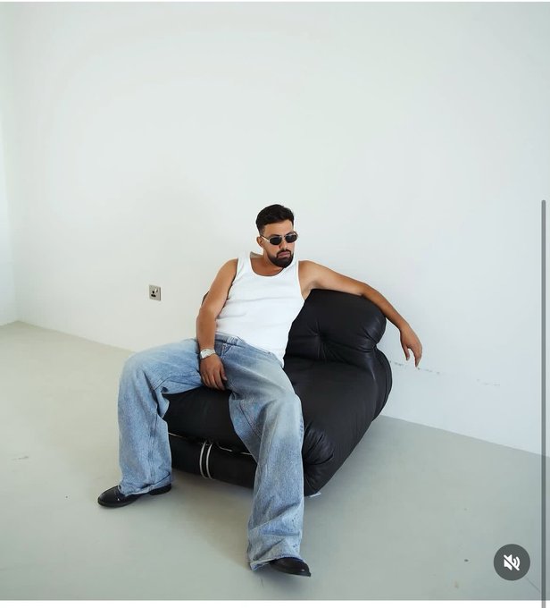 A stylish man with a beard and sunglasses wearing a white tank top, baggy blue jeans, and a watch, posing on a black leather chair in a minimalist white room.