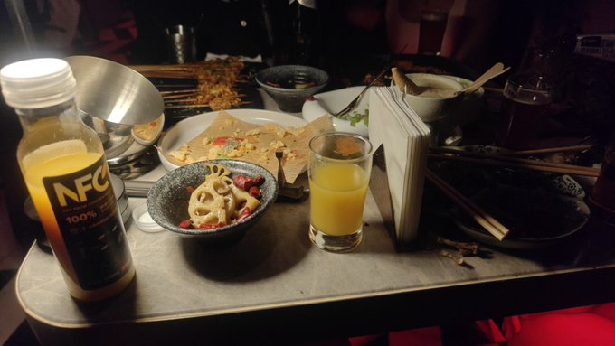 A bottle of NFC 100% orange juice sits alongside a glass of the same juice and various Asian-style dishes on a dimly lit restaurant table, suggesting the drink was ordered to accompany the meal. The caption's comment about something feeling "completely different" when touched likely refers to the distinctive, slightly thicker texture of this particular brand of NFC (Not From Concentrate) juice compared to typical reconstituted varieties.