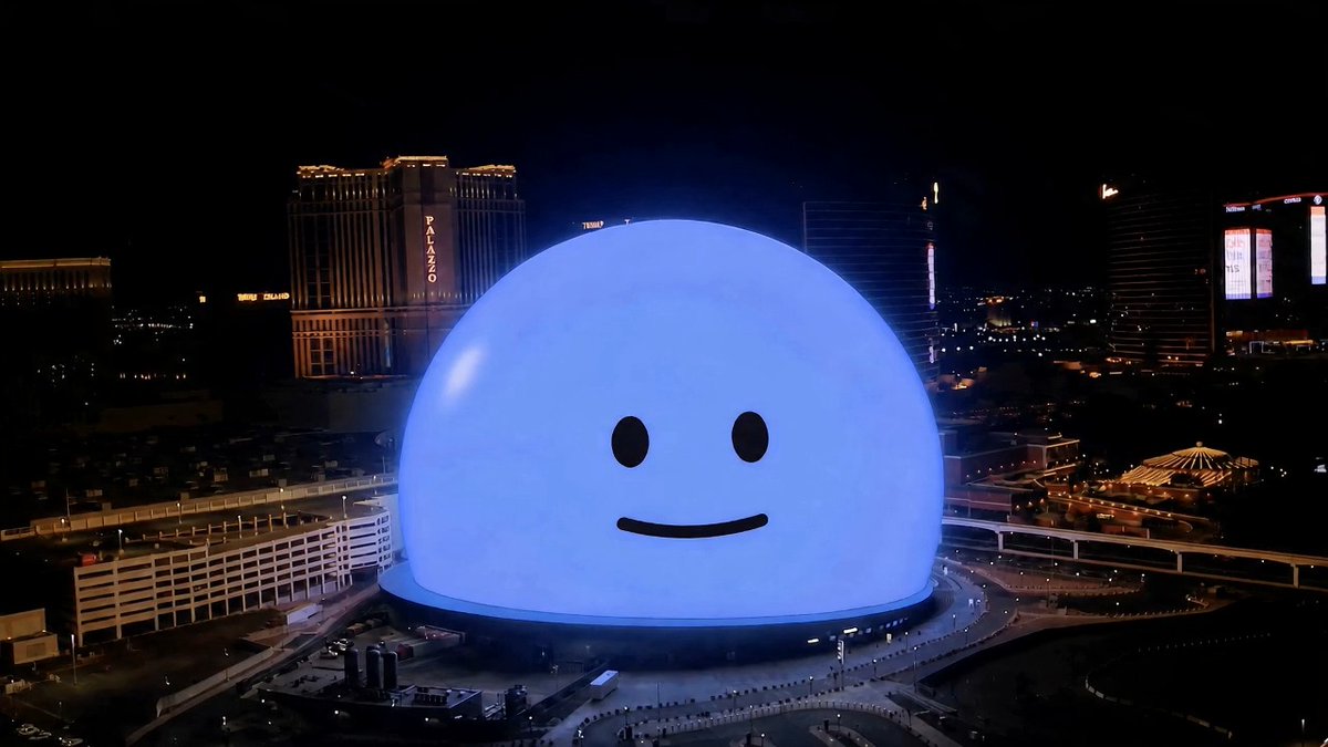 A giant illuminated blue spherical structure with a simple smiley face dominates the Las Vegas skyline at night, situated near The Palazzo and Treasure Island casinos with their illuminated signs visible in the background.