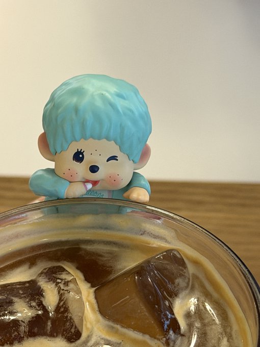 A small Monchhichi-style toy figure with blue hair and a winking expression peeks over the edge of a glass containing iced coffee.