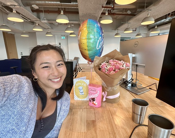 A smiling woman takes a selfie at her office desk, which is adorned with a rainbow "Congratulations" balloon, a bouquet of pink roses, and a "New Job" greeting card featuring a cactus illustration, celebrating her bittersweet last day at the Starknet Foundation after two years.