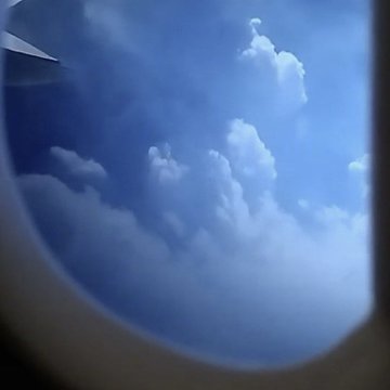 A view through an airplane window showing fluffy white clouds against a blue sky, with part of the airplane wing visible in the upper left corner.