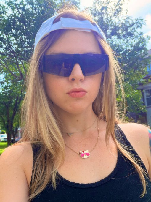 A selfie of a young woman with long blonde hair wearing dark rectangular sunglasses, a white visor, a black tank top, and layered gold necklaces with a pink pendant.