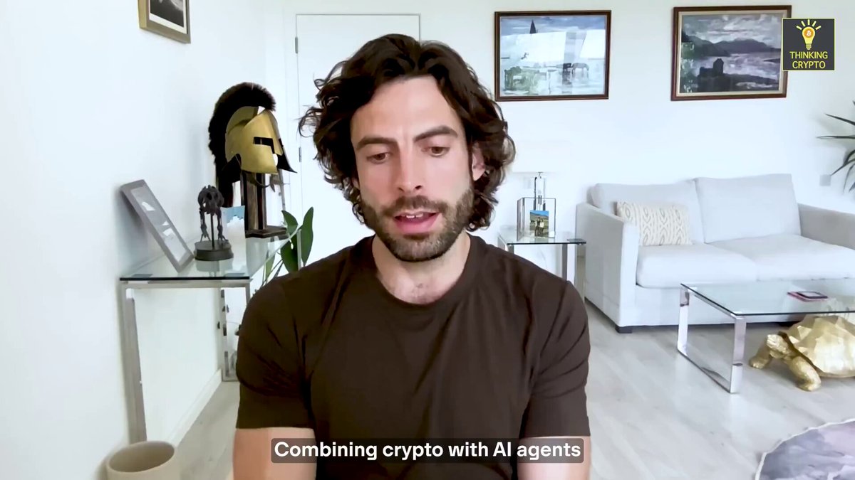The image shows a man sitting at a desk with a computer monitor in front of him. The background features a white wall, a couch, and several framed pictures hanging above the couch. There are also two potted plants placed near the couch. The main focus is on the man who appears to be talking about combining crypto with AI agents or robots.