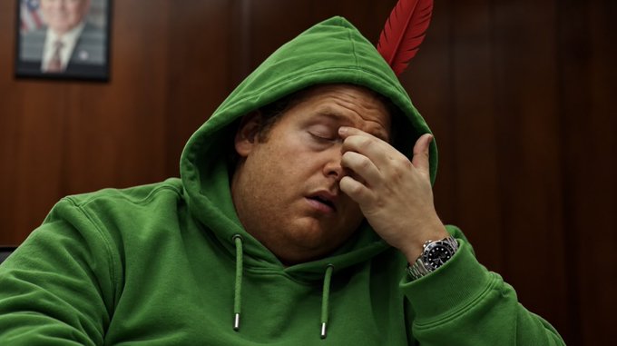 A man wearing a green hoodie with a red feather attached to it, rubbing his forehead with a tired expression, with a watch visible on his wrist.