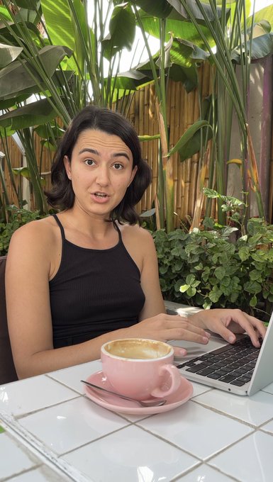 A woman in a black tank top sitting at a tiled outdoor table, typing on a laptop with a pink coffee cup beside her, with tropical plants and a bamboo fence in the background.