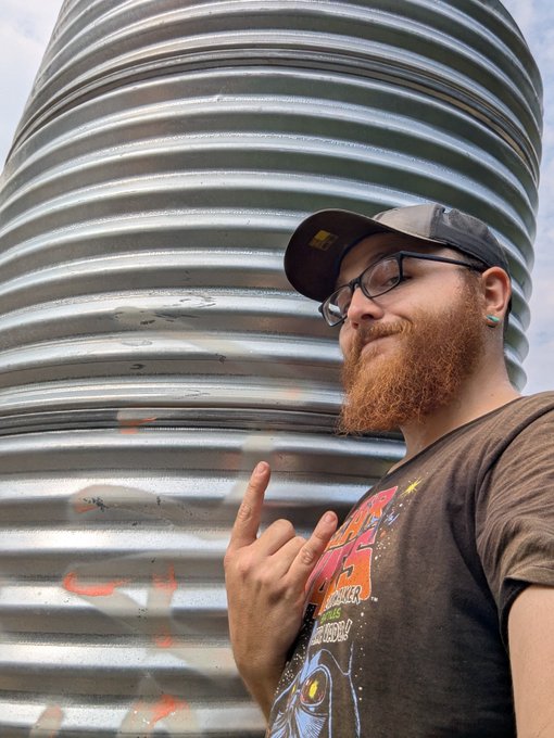 A man with a red beard, glasses, and a black cap stands next to a large corrugated metal silo, making a hand gesture while wearing a graphic t-shirt.