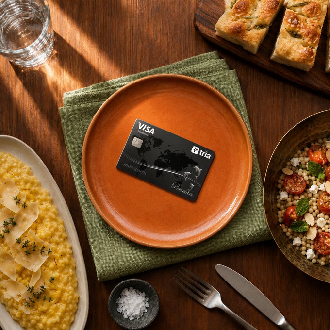 A black Visa card on a green napkin atop an orange plate, surrounded by dishes including risotto, focaccia bread, and a couscous salad on a wooden table.