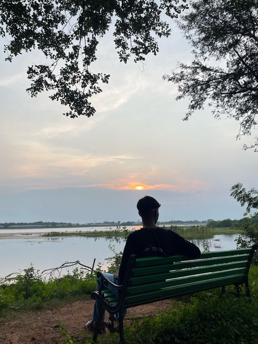 A person in a dark cap and shirt sitting on a green bench at sunset, looking out over a serene lake with trees framing the sky.
