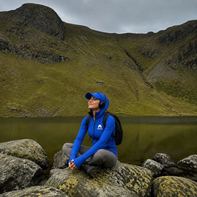 A woman wearing a blue hooded jacket and cap sits peacefully on a mossy rock by a mountain lake, with rugged green peaks rising behind her under an overcast sky.