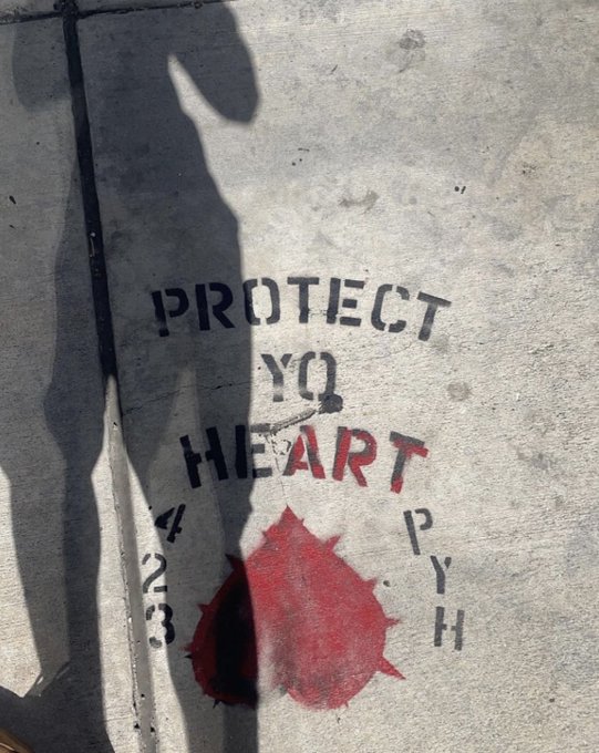 Stencil graffiti on pavement reading "PROTECT YQ HEART" with "423 PYH" and a red heart symbol, with shadows of two people visible.