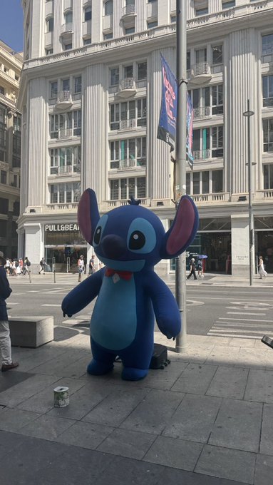 A person in a full-body Stitch costume standing on a city sidewalk in front of a Pull&Bear store with a stone building in the background.