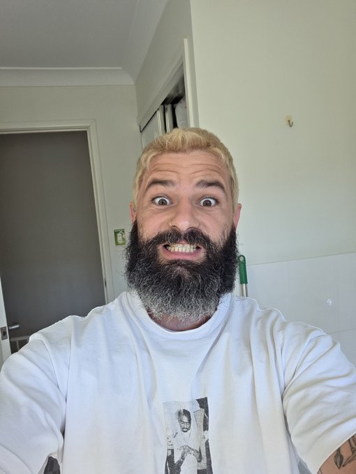 A selfie of a man with blonde hair, a thick dark beard with grey patches, and a wide-eyed expression, wearing a white t-shirt with a graphic print in a room with white walls and a doorway behind him.