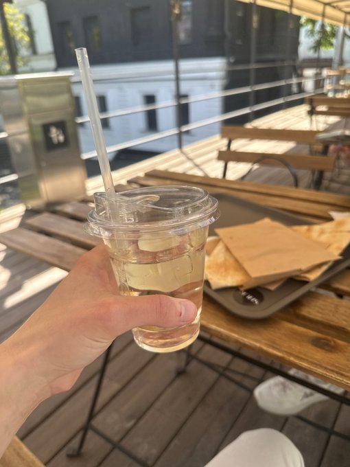 A hand holds a clear plastic cup with a straw containing a light-colored beverage at an outdoor wooden table setting, with napkins and a menu visible nearby.