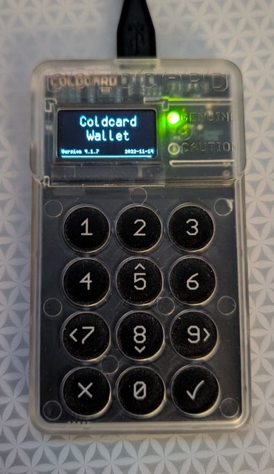 A Coldcard hardware wallet with a transparent case displaying 'Coldcard Wallet' on its screen, version 'v.1.7.7', dated '2023-11-14', with a numeric keypad.