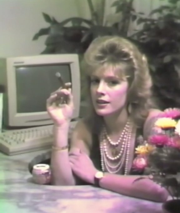 The image features a woman sitting at a desk with an old computer monitor, keyboard, and mouse. She has her arm resting on the desk while holding a cigarette. The background includes a potted plant and flowers, adding a touch of nature to the scene. The photo is accompanied by text that reads "Another afternoon lull at the office of the future 8 editions: objkt.com/tokens/KT1VyZg". This suggests that the image might be related to an article or post about the woman's work environment, possibly discussing her experiences in a vintage setting with old technology and a more relaxed atmosphere compared to modern offices.
