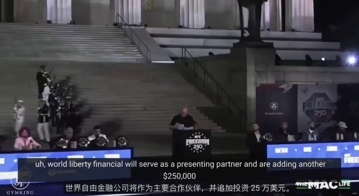 In this image, there's an event taking place where people are gathered at a stage, possibly for a presentation or a speech. One of the individuals standing on the stage appears to be speaking into a microphone. The setting includes a staircase behind them, adding to the atmosphere of the event. There is also a TV screen present in the scene which displays some text that reads "uah world liberty financial will serve as a presenting partner and are adding another $1,500,000". This could be referring to a partnership or collaboration between two companies with different strengths or specialties. The audience seems engaged in what is being discussed at the event, potentially providing feedback on this new partnership opportunity.