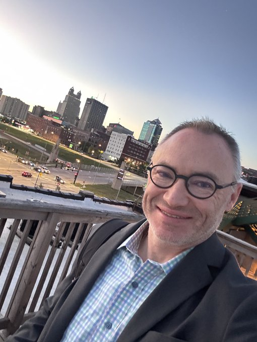 A middle-aged man with glasses and short hair takes a selfie on a bridge or elevated walkway, wearing a dark jacket over a plaid shirt, with a city skyline and sunset visible in the background.
