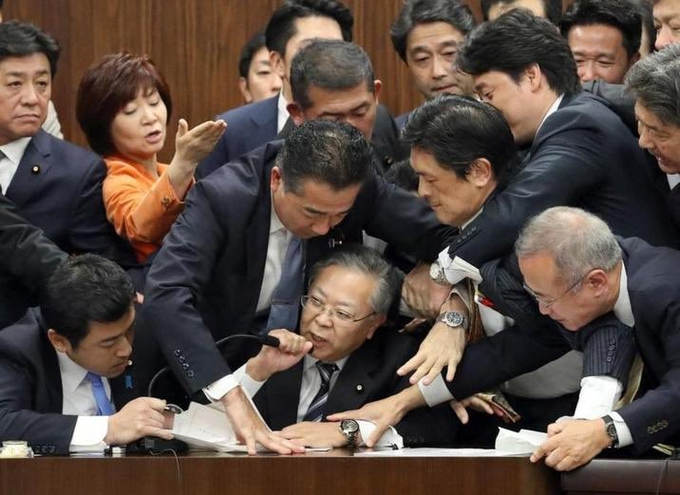 A chaotic scene of Japanese politicians in suits crowding around a desk during what appears to be a parliamentary confrontation.