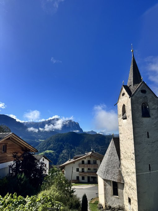 A picturesque view shows a tall stone church with a pointed steeple in the foreground, overlooking traditional alpine houses with a large, misty mountain peak visible in the background under a bright blue sky.